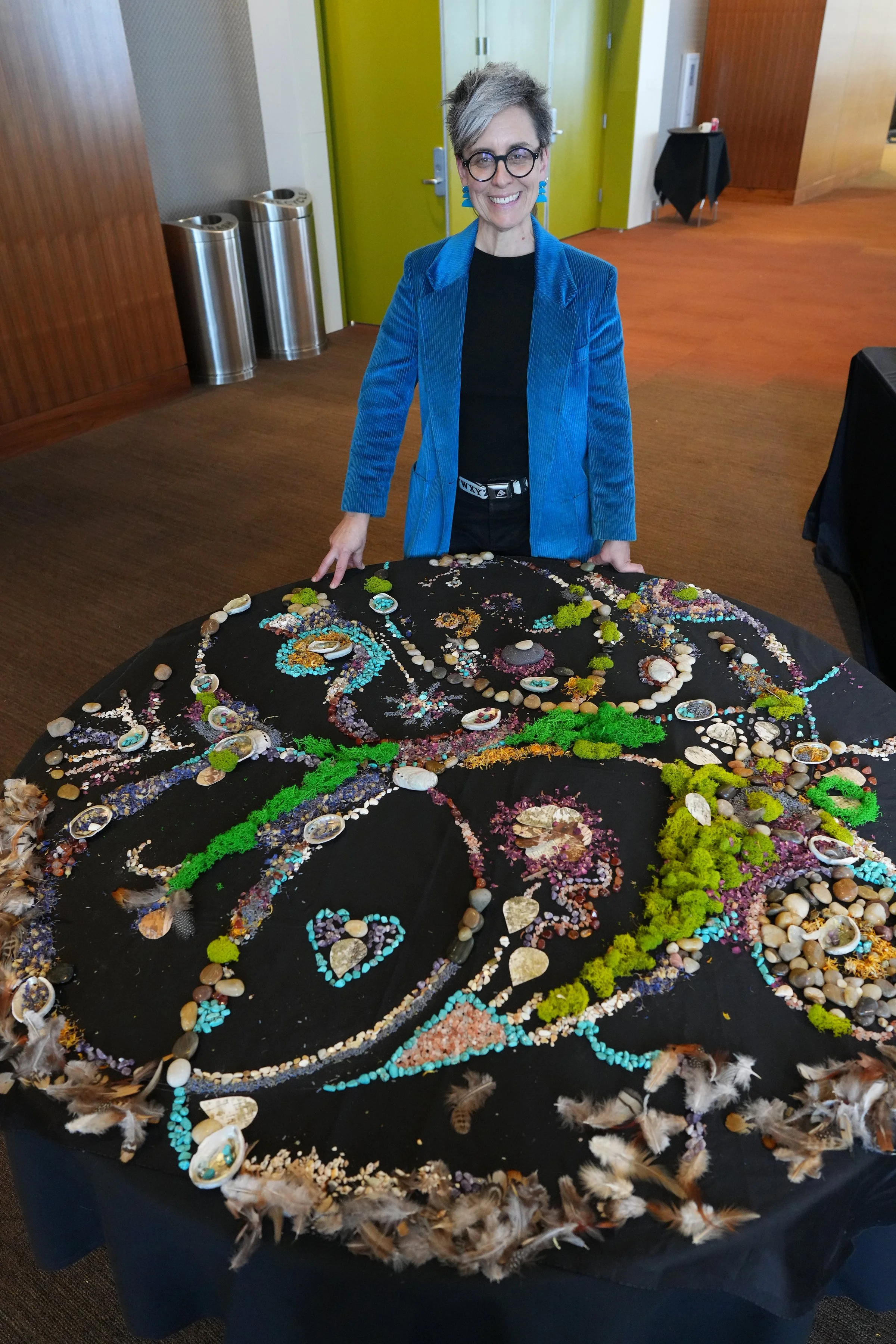 A woman is standing by a round table covered in black linen and decorated with different objects with different textures, sizes, colors as an art table.
