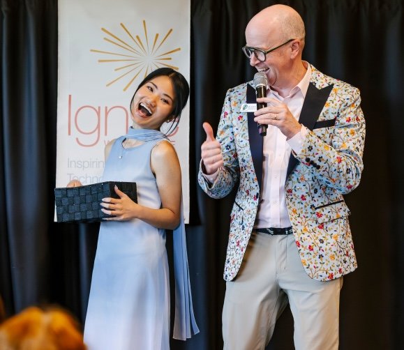 A woman is holding a box onstage while a man with a microhpone has his thumb up in approval.