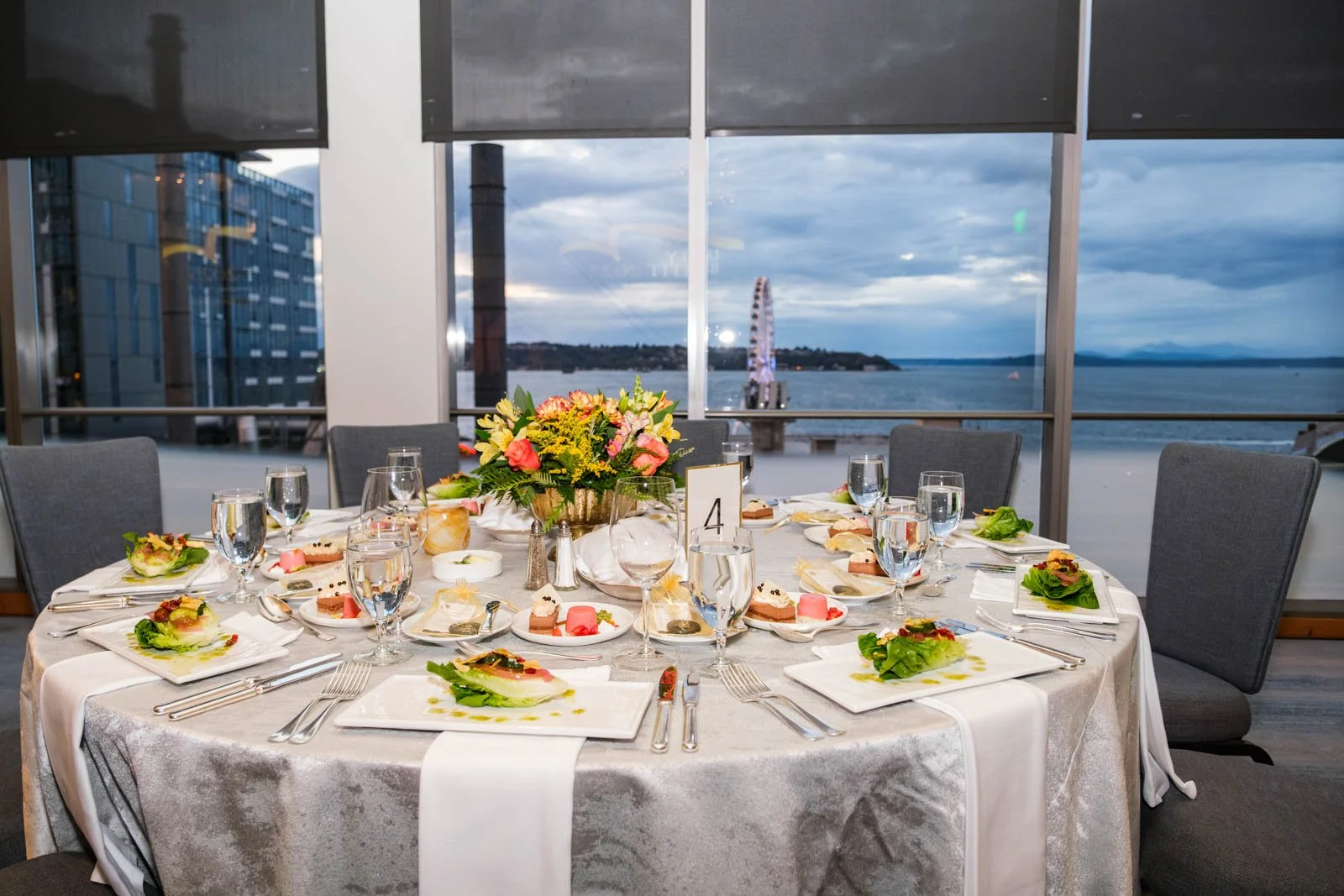 A round table with pre-set salads served with dessert, while the bayside view at dusk is seen through the window's view.