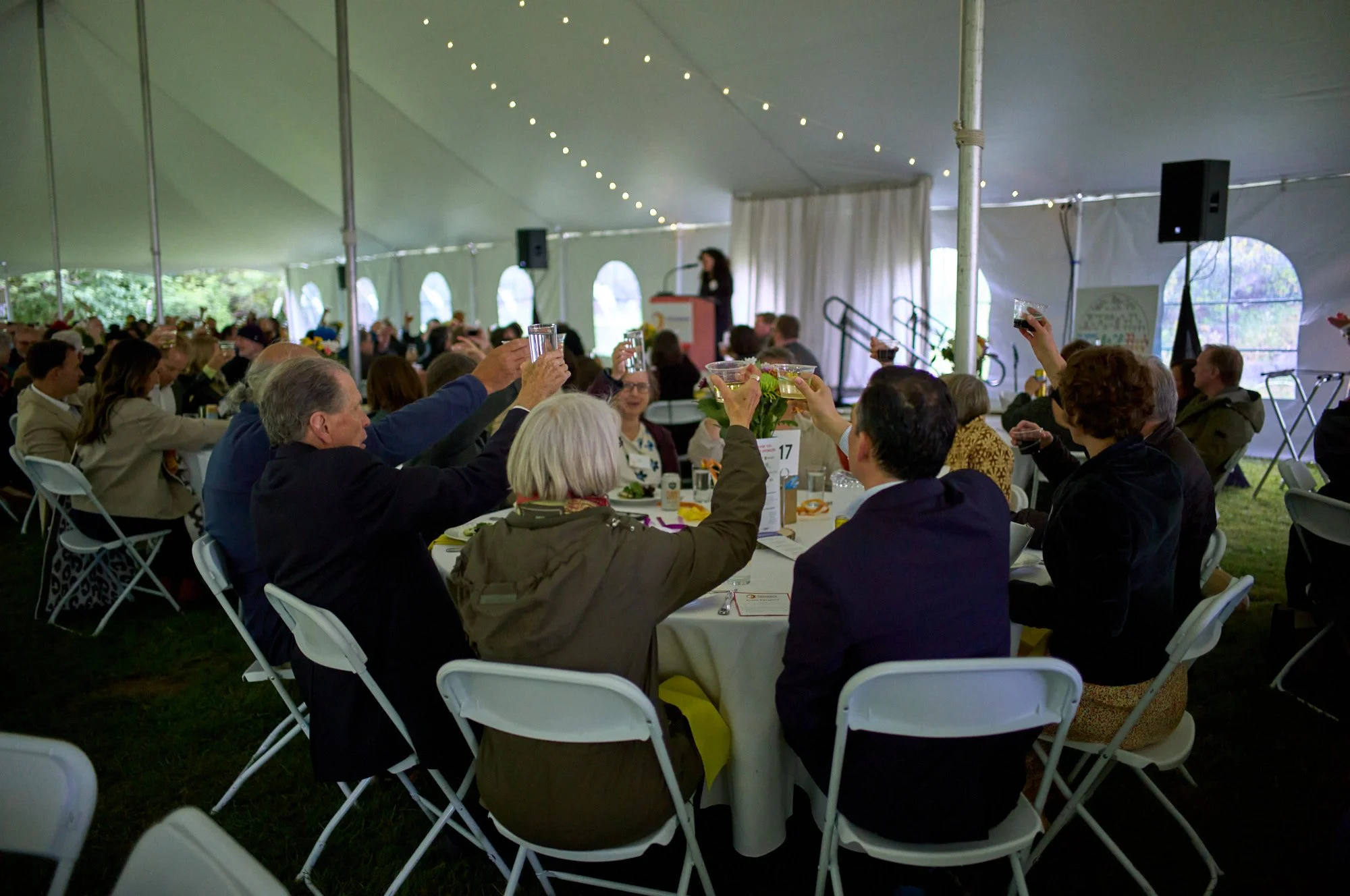 Guests on a round table are seen toasting in the foreground while there is a speaker on the podium on a stage in the background.