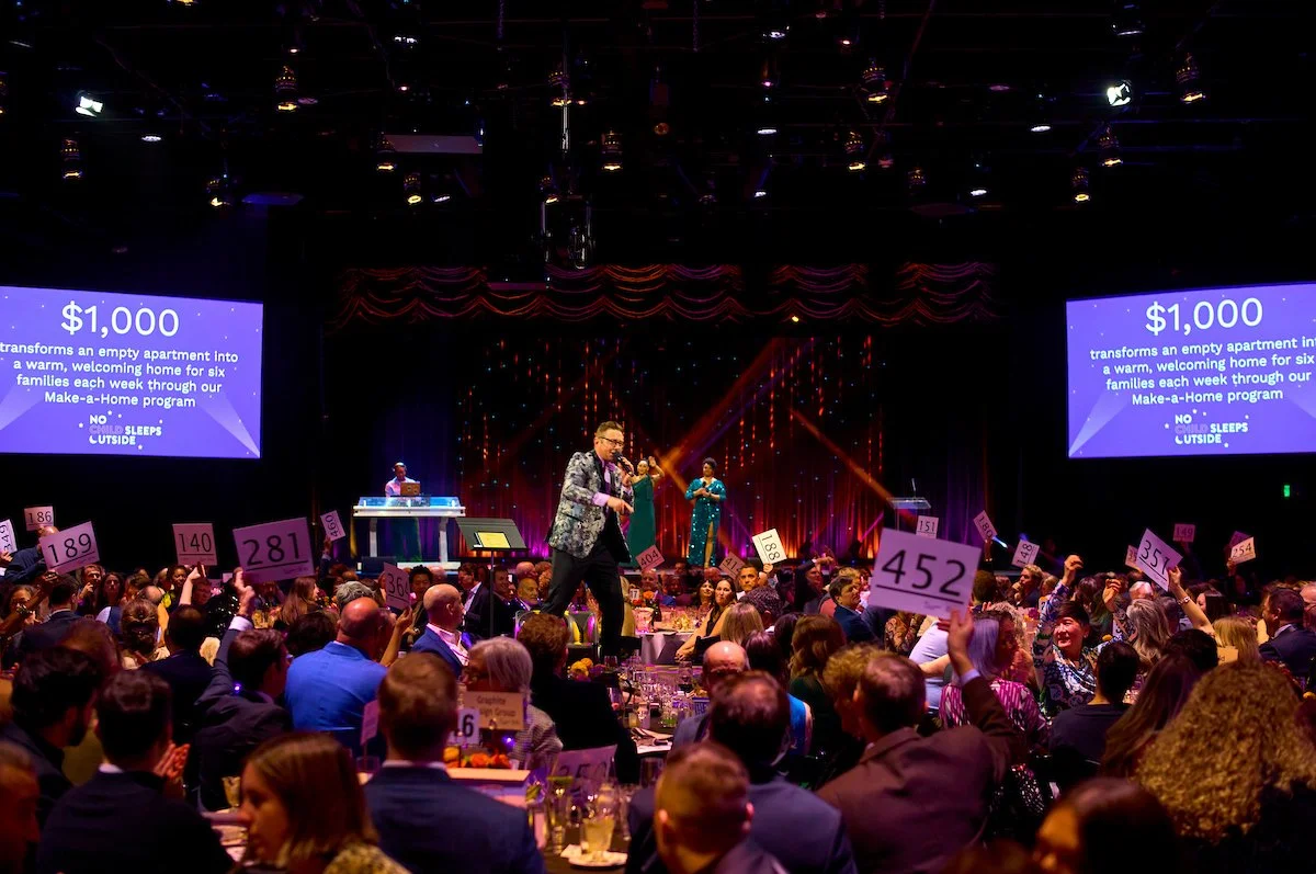 A host reads bid numbers in the middle of guests seated on round tables with seated guests in formal wear.
