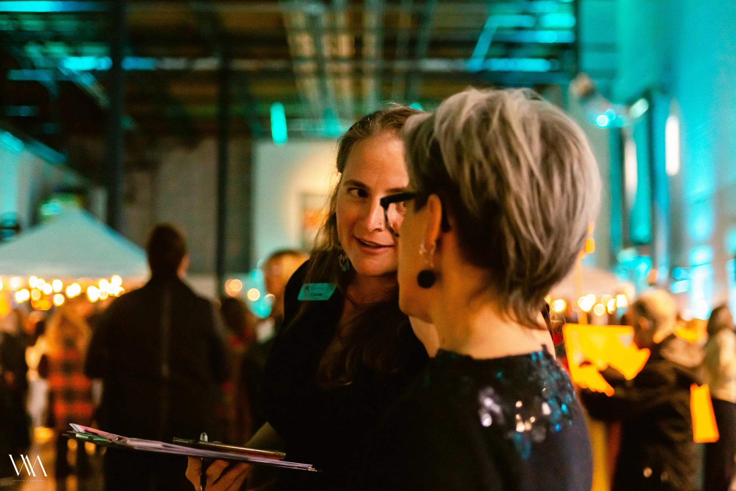 Two women at an event, are discussing and facing each other, one has her phone and clipboard on hand..