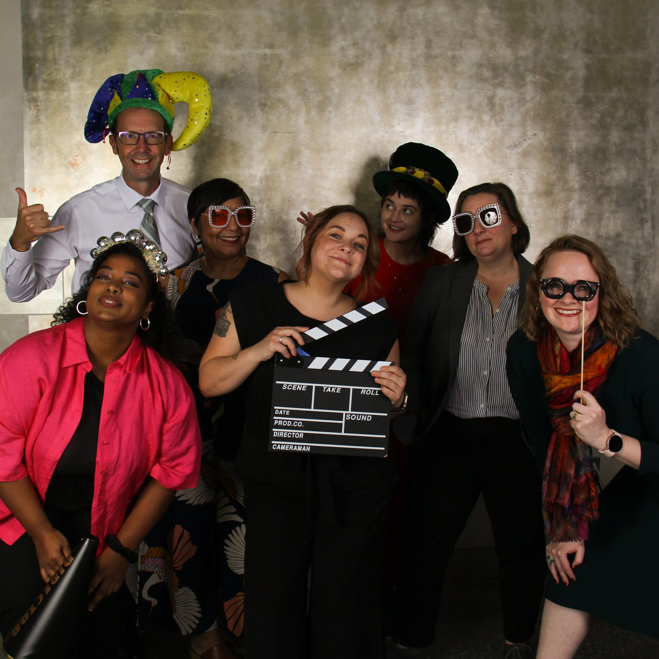 A group of seven individuals hams it up for the camera, holding silly props, wearing sparkly hats and sunglasses.