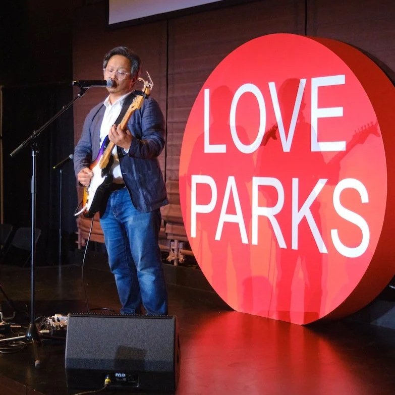 A man in jeans and a jacket plays a guitar, next to a large sign reading "Love Parks".