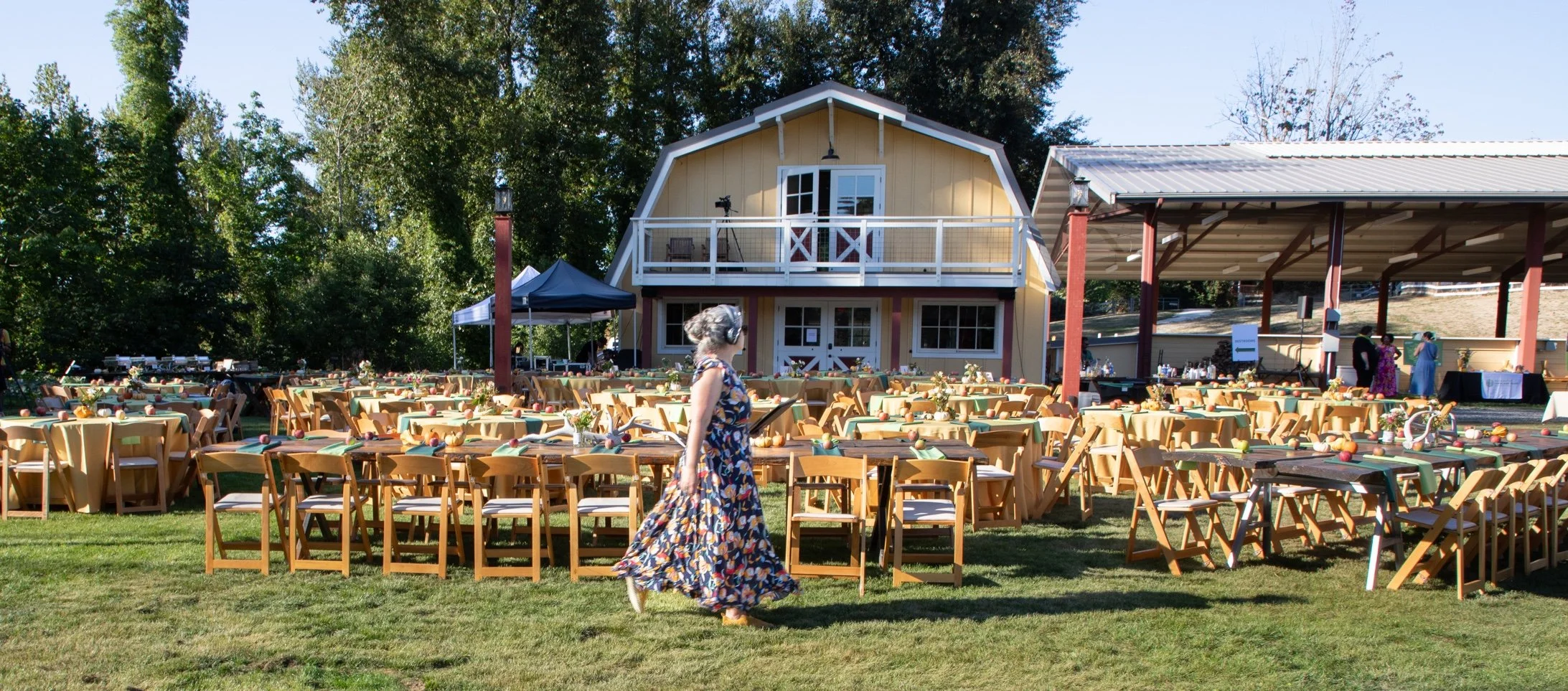 An event planner in a flowing sundress carries her clipboard as she walks past a beautiful party set up on a lawn, in front of a barn.