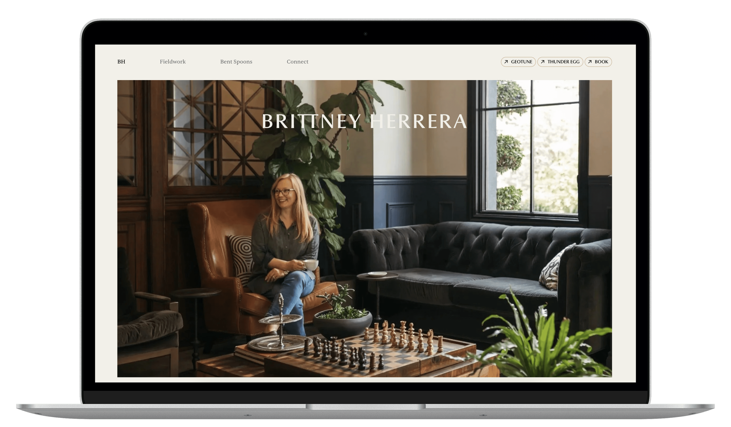 Laptop displaying a website with a woman sitting on a brown leather chair in a cozy, well-decorated room with large windows, a black tufted sofa, plants, and a chessboard on a wooden table.
