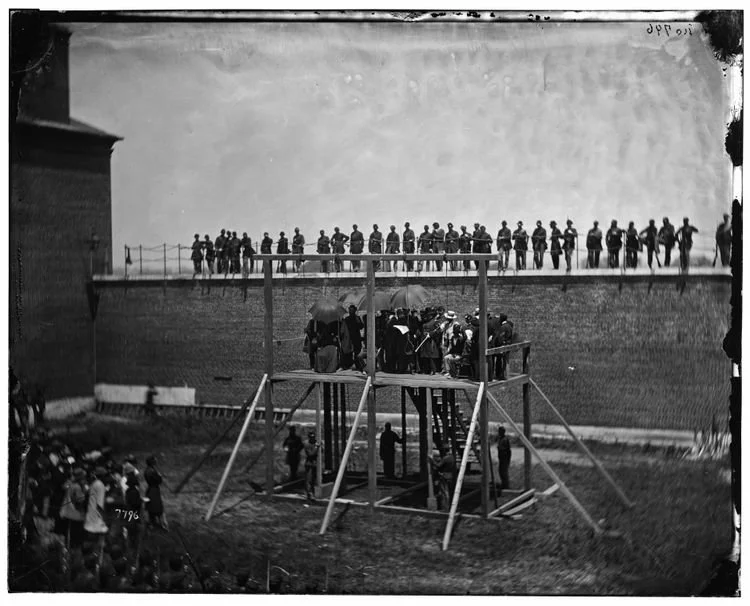 The gallows with executioners standing below and a crowd gathered to watch.
