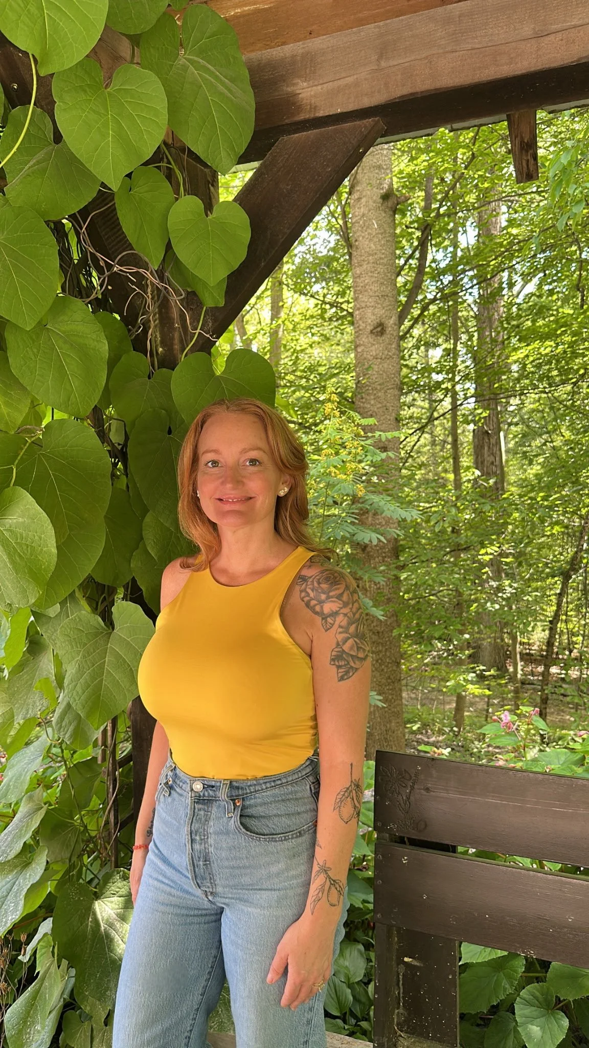 A woman with red hair and tattoos wearing a yellow sleeveless top and jeans, standing outdoors among trees and large green leaves, smiling at the camera.