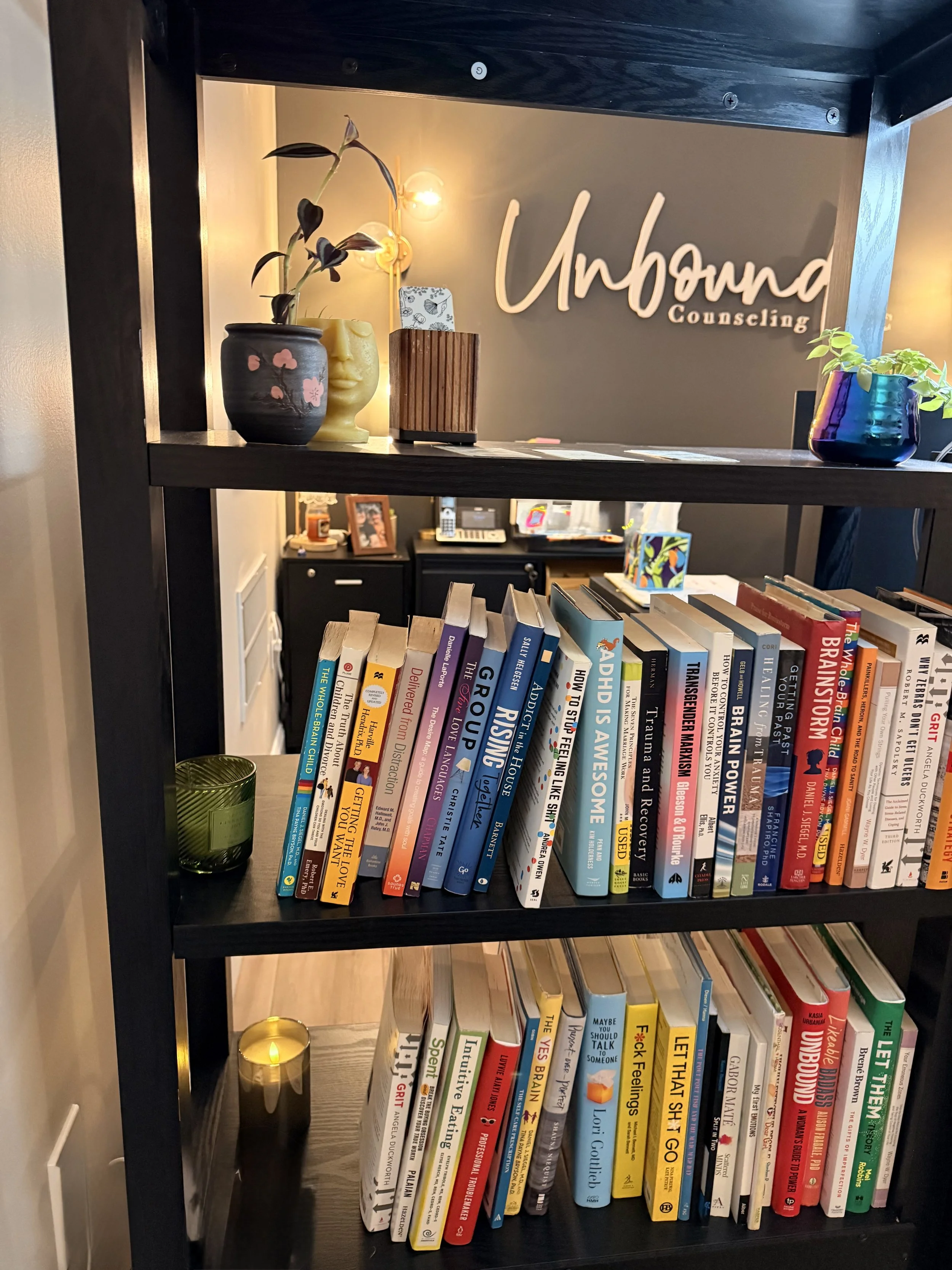 Bookshelf filled with colorful books, a green candle holder at the bottom left, decorative vases with plants and a face sculpture, a sign reading 'Unbound Counseling' on the wall, cozy lamps in the background, and the room appears to be a counseling office.
