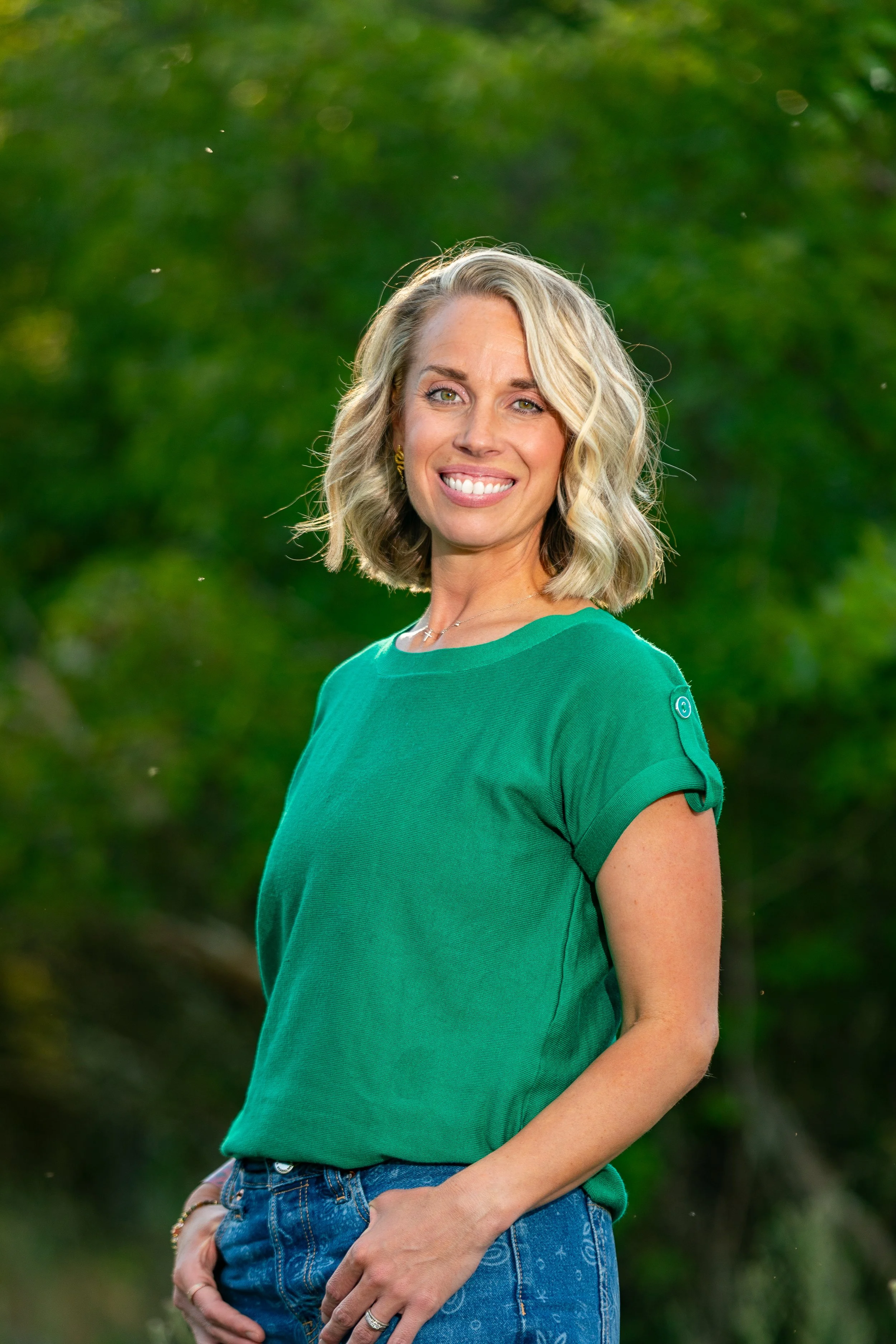 A woman with shoulder-length blonde hair smiling outdoors, wearing a green short-sleeve T-shirt and blue jeans, with a lush green background.