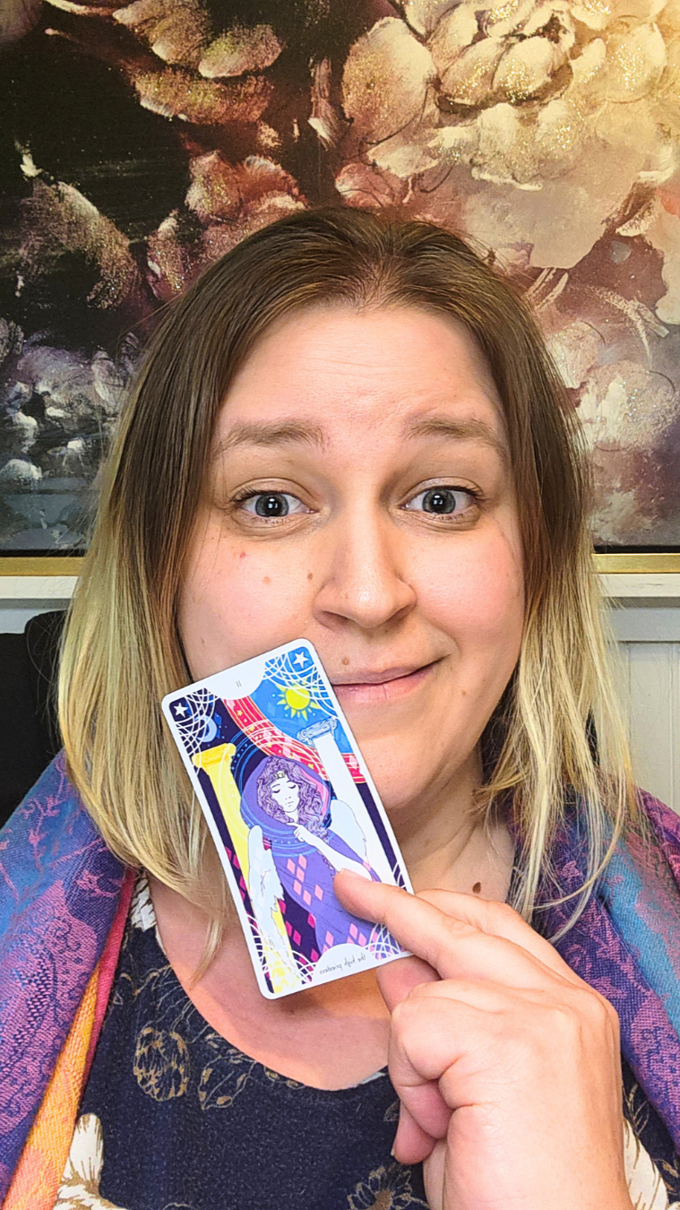A person holding a tarot card in front of her face, smiling for the camera, with a large floral painting in the background.