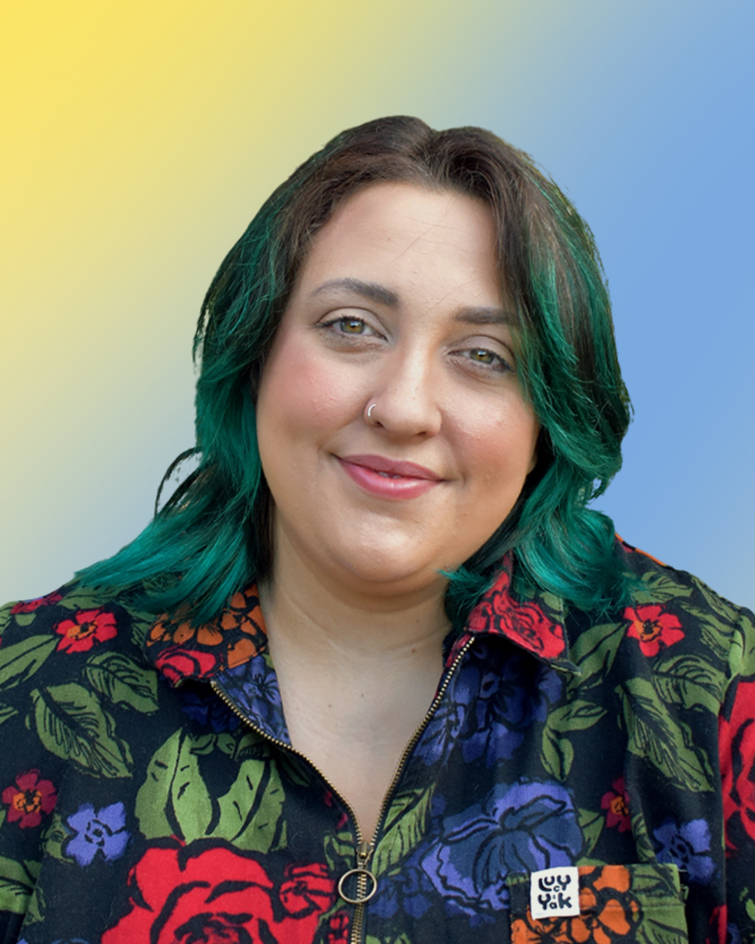 Portrait of a non-binary person with shoulder-length hair dyed green and black, wearing a floral jacket with a zipper, against a blurred yellow and blue background.