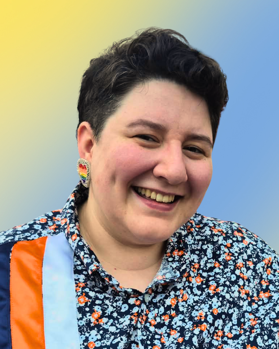 A smiling person with dark hair wearing a beaded rainbow heart earring and paisley shirt with ribbons