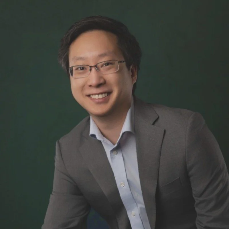 A young man in a gray suit and glasses smiling against a green background.