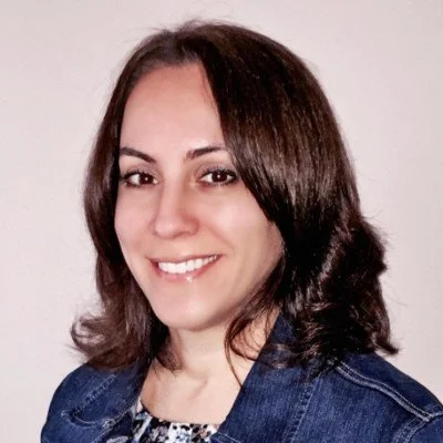 A woman with shoulder-length brown hair smiling at the camera, wearing a denim jacket over a patterned top, against a plain light background.