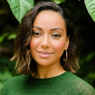 A woman with shoulder-length wavy hair wearing a green sweater and hoop earrings, smiling outdoors with green foliage in the background.