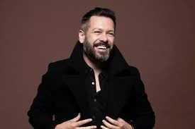 A man with a beard and short hair, smiling, wearing a black jacket against a brown background.