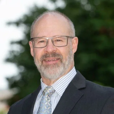 Portrait of a middle-aged man with glasses, a beard, wearing a suit and tie, outdoors with trees in the background.