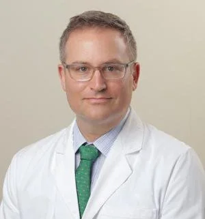 Professional portrait of a male doctor wearing glasses, a white coat, a light blue shirt, and a green tie, standing against a plain beige background.
