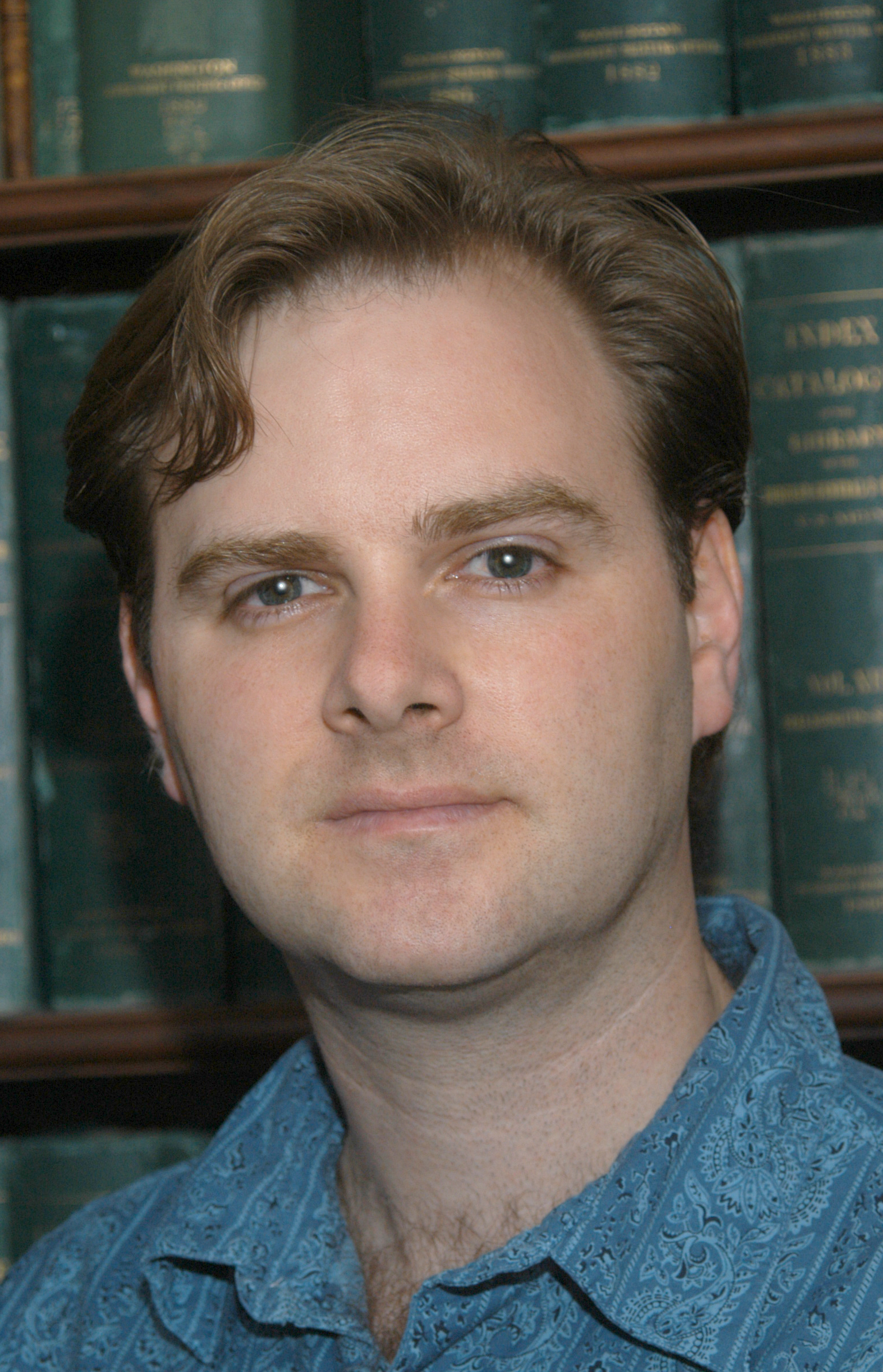 A close-up portrait of a man with light skin, brown hair, and blue eyes, standing in front of a bookshelf filled with thick, green law books.