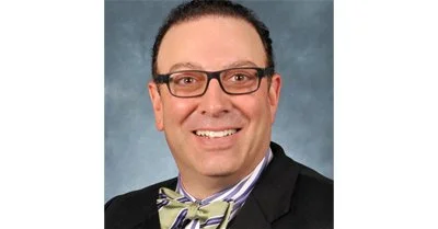 A man with glasses and dark hair, smiling, dressed in a black suit with a purple and white striped shirt and a yellow bow tie.