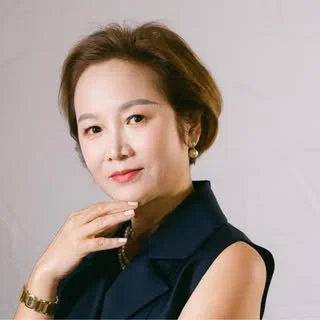 Portrait of a woman with short hair wearing a dark sleeveless top and gold jewelry, posing with her hand near her face against a plain background.