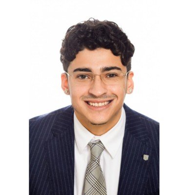 Portrait of a young man with curly dark hair wearing glasses, a dark pinstripe suit, white shirt, and patterned tie, smiling against a plain white background.