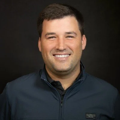 A smiling man with short dark hair, wearing a navy blue jacket, against a dark background.