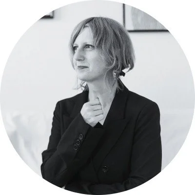 A woman with shoulder-length hair wearing a blazer, sitting with her hand on her chin in a thoughtful pose.
