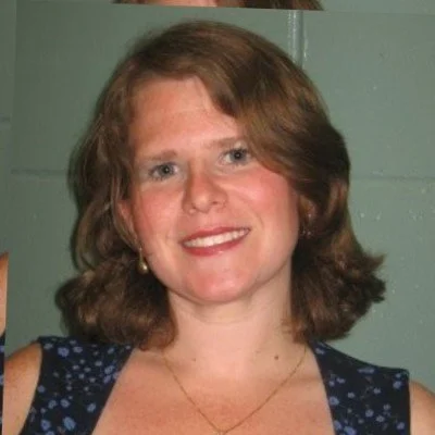 A woman with shoulder-length brown hair smiling at the camera, wearing a blue dress with white polka dots and gold earrings.