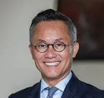 A professional man with glasses, short dark hair, wearing a dark suit and blue shirt, smiling in an indoor setting.