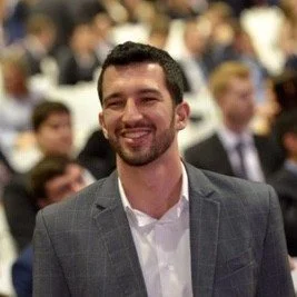 A smiling man in a gray suit at a gathering or conference, with a blurred crowd in the background.