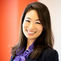 Portrait of a smiling woman with long dark hair wearing a purple blouse against an orange background.
