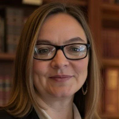 Close-up of a woman with shoulder-length brown hair, wearing glasses, smiling in front of a bookshelf.