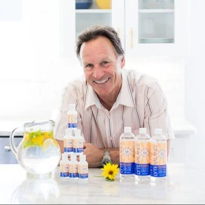 Smiling man sitting at a white table with bottles of skincare products, a jar of lemon water, and a yellow flower in a bright, modern kitchen.