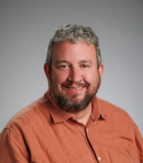 A smiling man with short gray hair and a beard, wearing an orange button-up shirt against a plain gray background.
