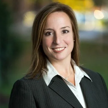 A woman with brown hair smiling outdoors, wearing a black blazer and white shirt.