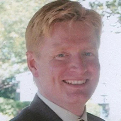 A smiling young man with blonde hair, wearing a suit, outdoors with trees in the background.