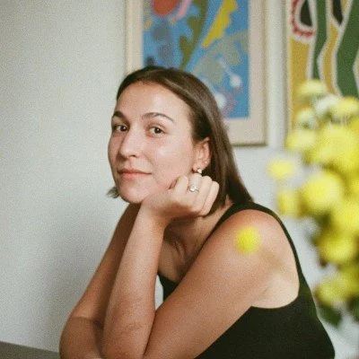 A woman with short brown hair resting her chin on her hand, sitting indoors with colorful artwork on the wall behind her.