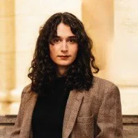 A woman with dark curly hair wearing a black top and brown blazer, standing indoors.