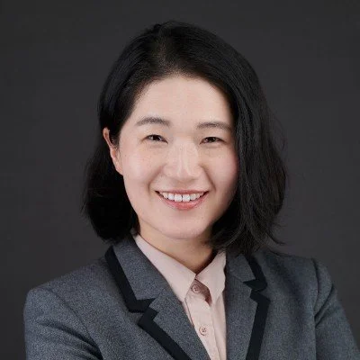 Professional woman with shoulder-length black hair, smiling, wearing a gray blazer and a light-colored shirt, against a dark background.