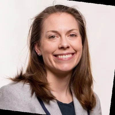 A smiling woman with shoulder-length brown hair, wearing a gray blazer and a dark top, against a gray background.