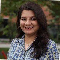 Portrait of a smiling woman with long dark hair wearing a blue patterned shirt outdoors.