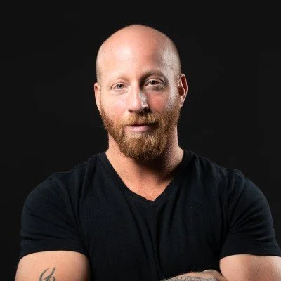 A man with a bald head and a beard, wearing a black T-shirt, posing against a black background.