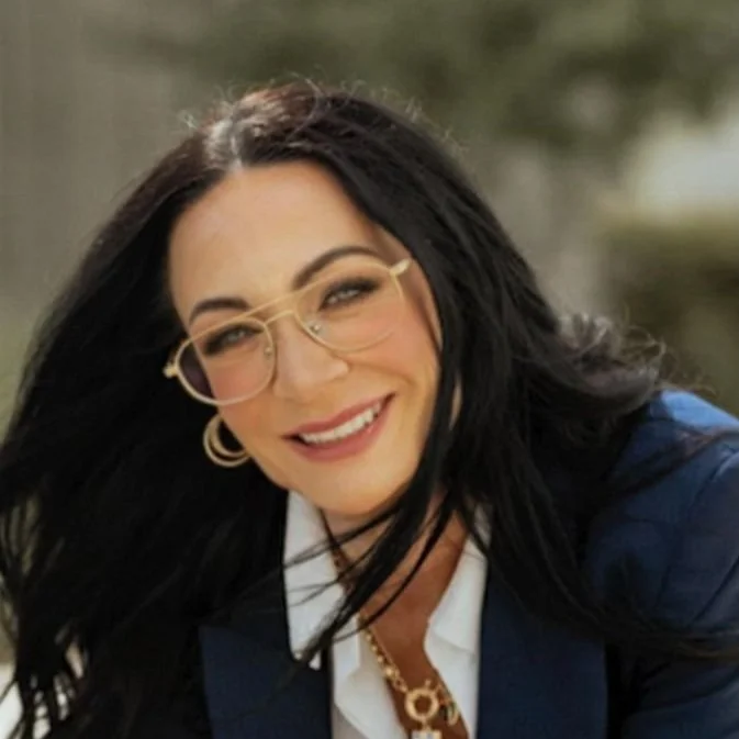 Smiling woman with black hair, glasses, and gold jewelry, wearing a blazer and white shirt.