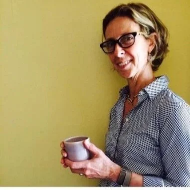 A woman with glasses holding a mug and smiling, standing against a yellow wall.