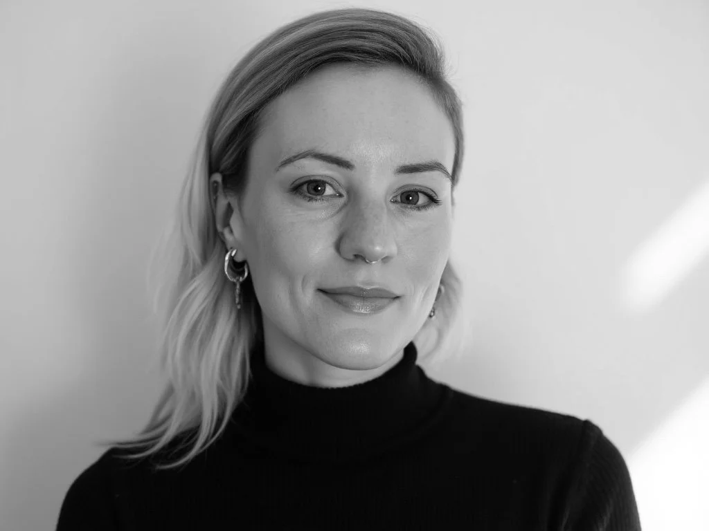 A black and white portrait of a woman with short, wavy blonde hair, wearing hoop earrings and a black turtleneck, smiling slightly against a plain background.