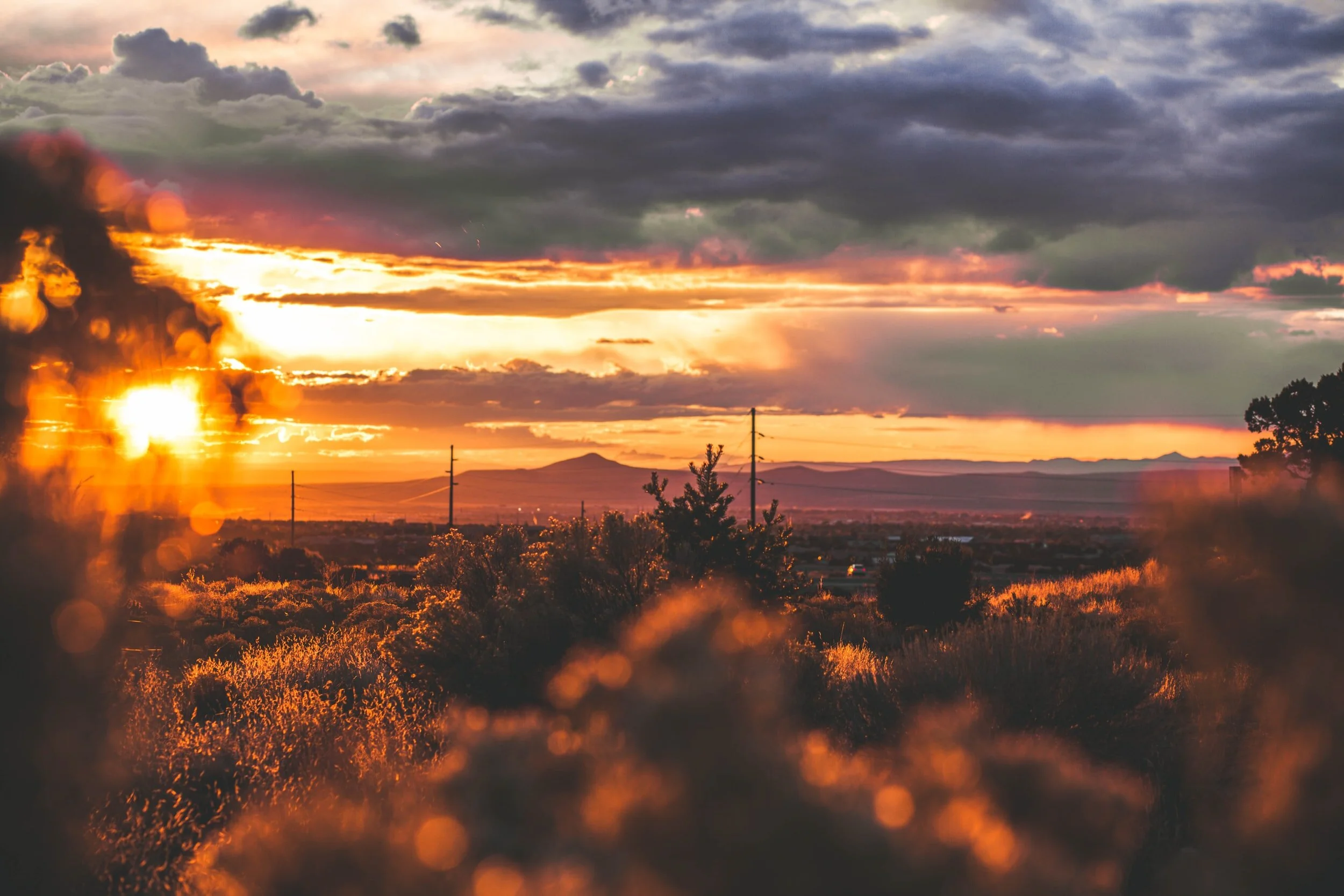 Sunset over a landscape with trees, hills, and power lines under a partly cloudy sky.