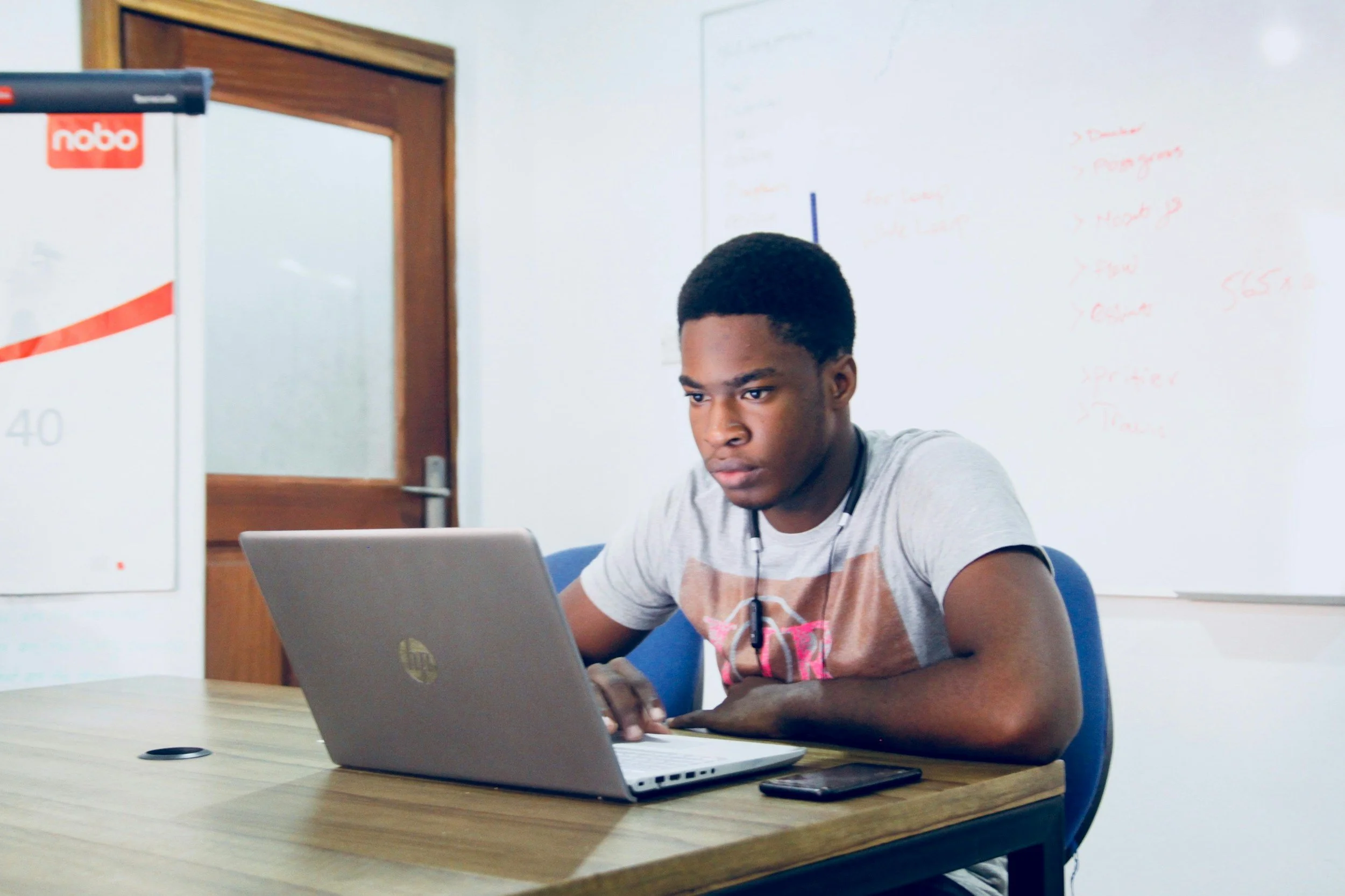 A young man working on a laptop at a desk in a classroom or office, with a smartphone and whiteboard in the background.