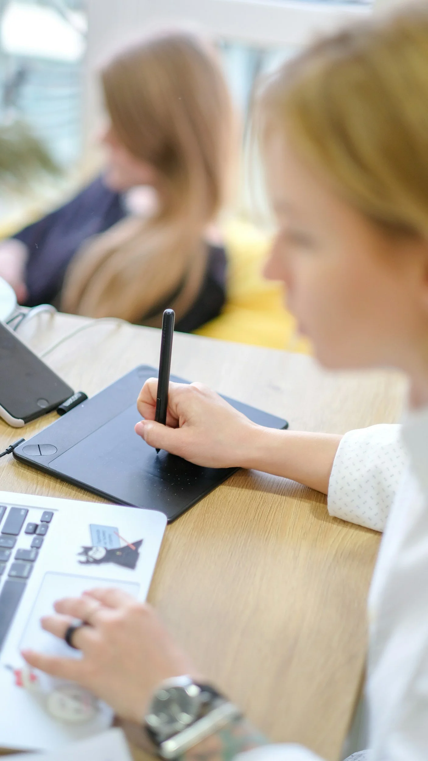 A woman using a graphics tablet and stylus at a desk with a laptop and smartphone nearby, while two women sit in the background.